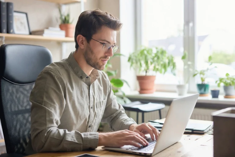 Fair-skinned man centered at desk typing on laptop in bright modern office